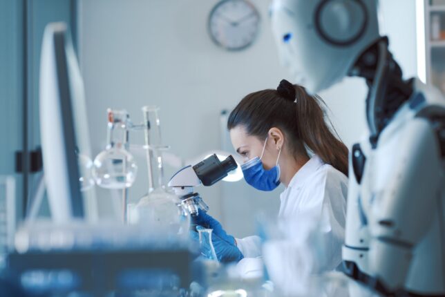 A lab scientist works at a microscope while, in the foreground, an anthropomorphic robot also works at a microscope, illustrating the idea of Industry 5.0