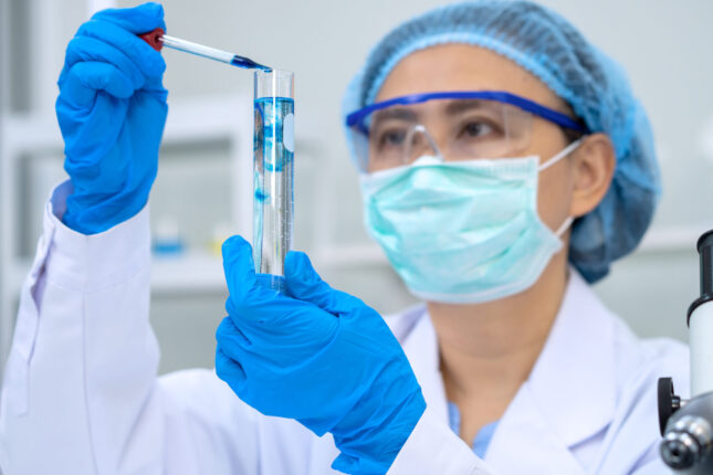 A QC technician in laboratory clothing pipettes solution into a test tube as part of standard quality operations