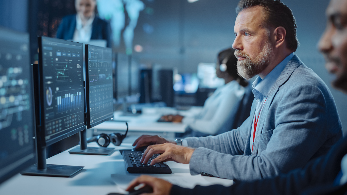 Portrait of Serious Professional Technical Controller Sitting at His Desk with Multiple Computer Displays Before Him. In the Background His Colleagues Working in System Control and Monitoring Center.