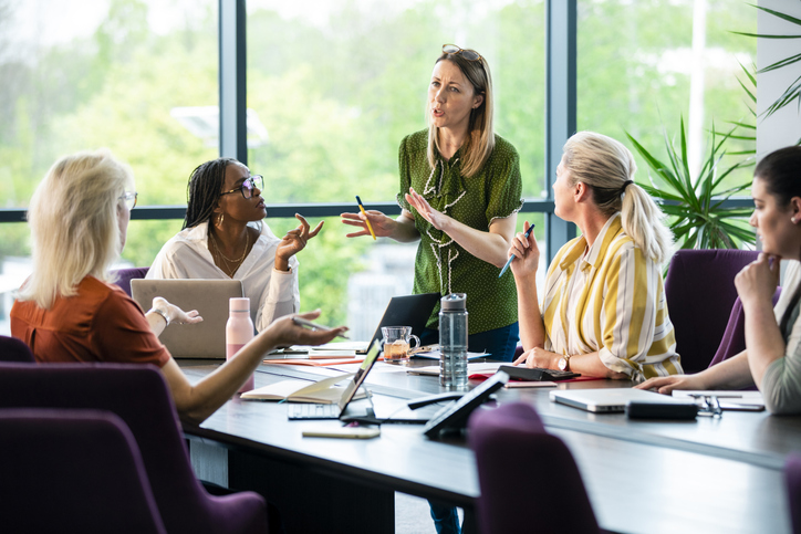 A wide-angle view of a board room meeting which is all women. They are discussing ideas and working on things together to come up with business opportunities.