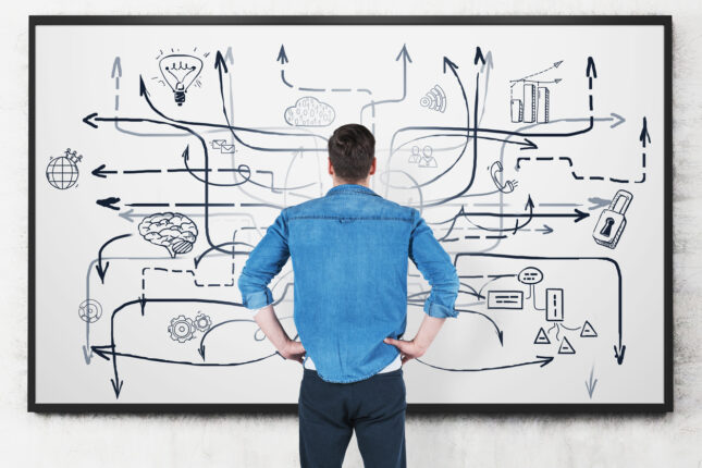 Rear view of a man looking in confusion at a whiteboard with a messy and complex diagram that could easily represent the complexity of QC operations