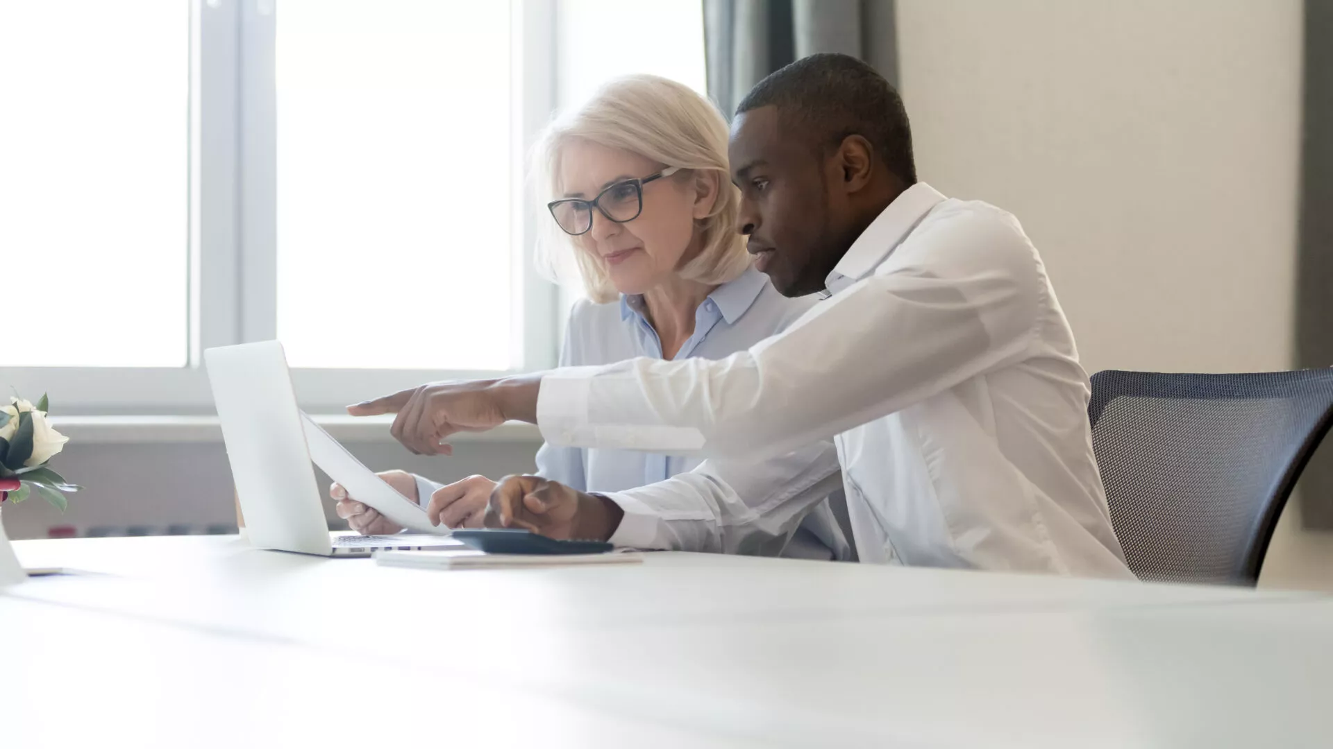 African american employee pointing at laptop discussing paperwork with colleague working together focused on computer task, black business employee helping giving online presentation to client
