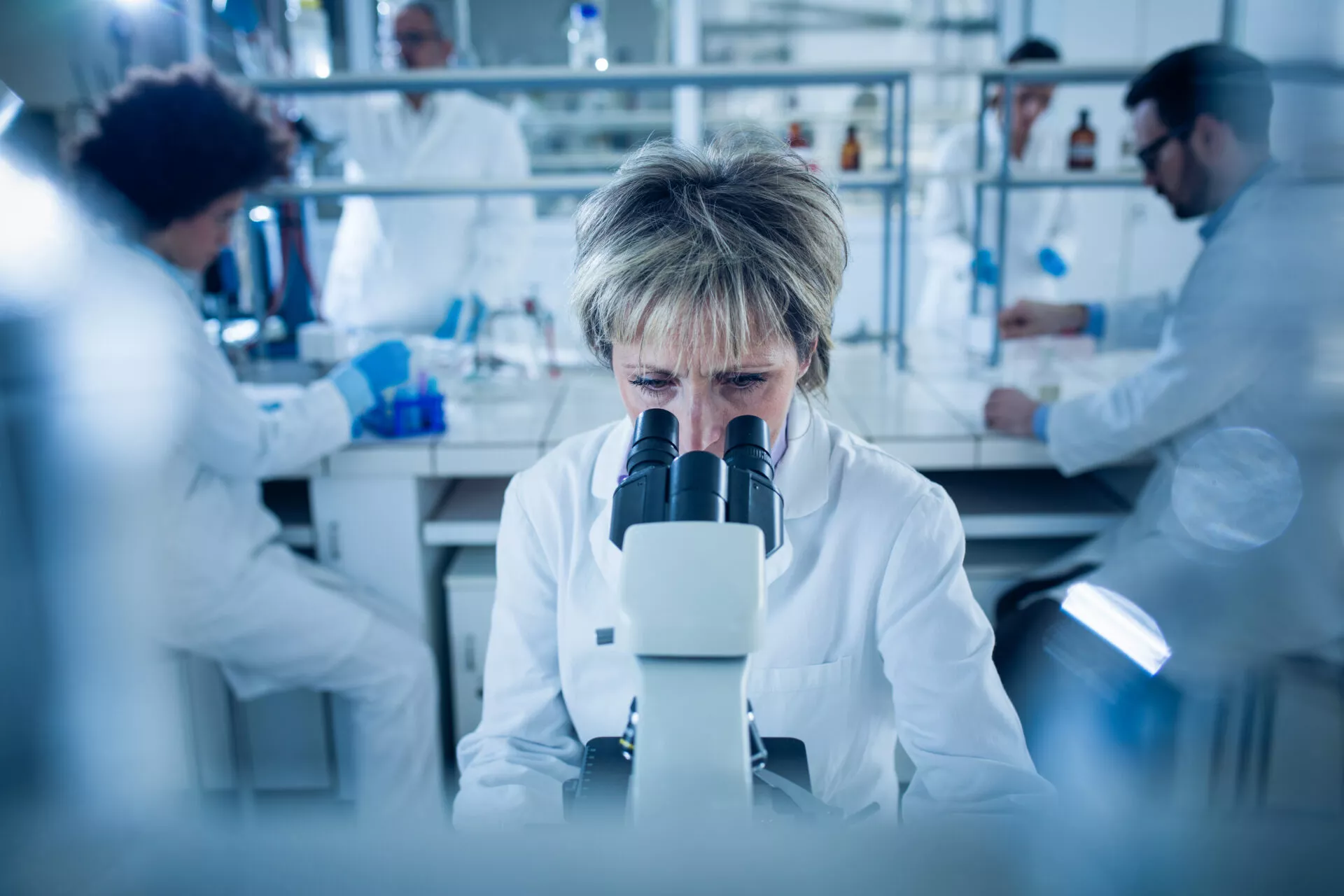 Senior forensic scientist analyzing something through a microscope in laboratory. There are people in the background.