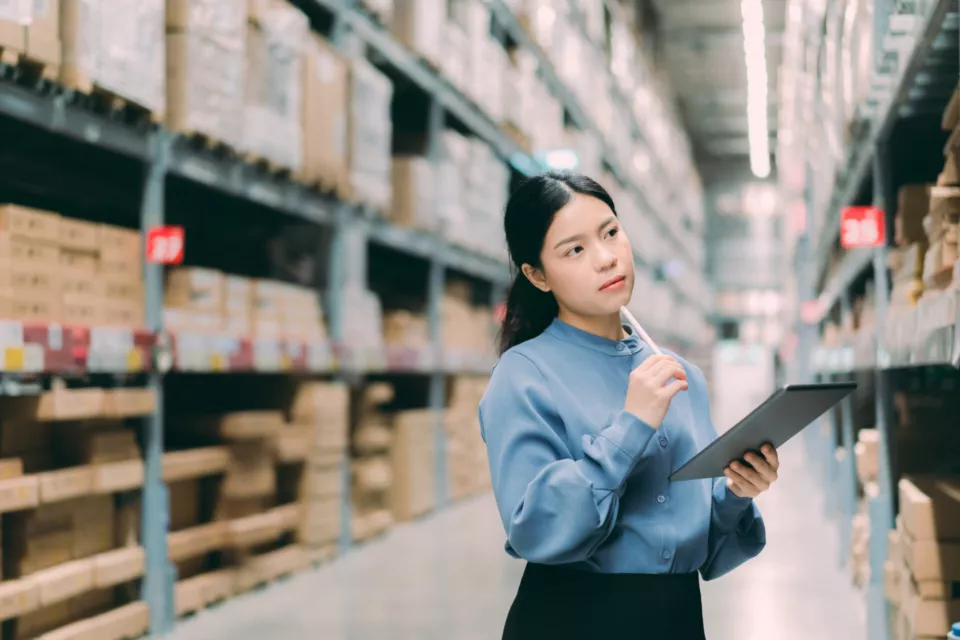 Young smart Asian business working woman thinking while using digital tablet to check goods on shelves for product management  in warehouse, Logistics business planning concept with copy space