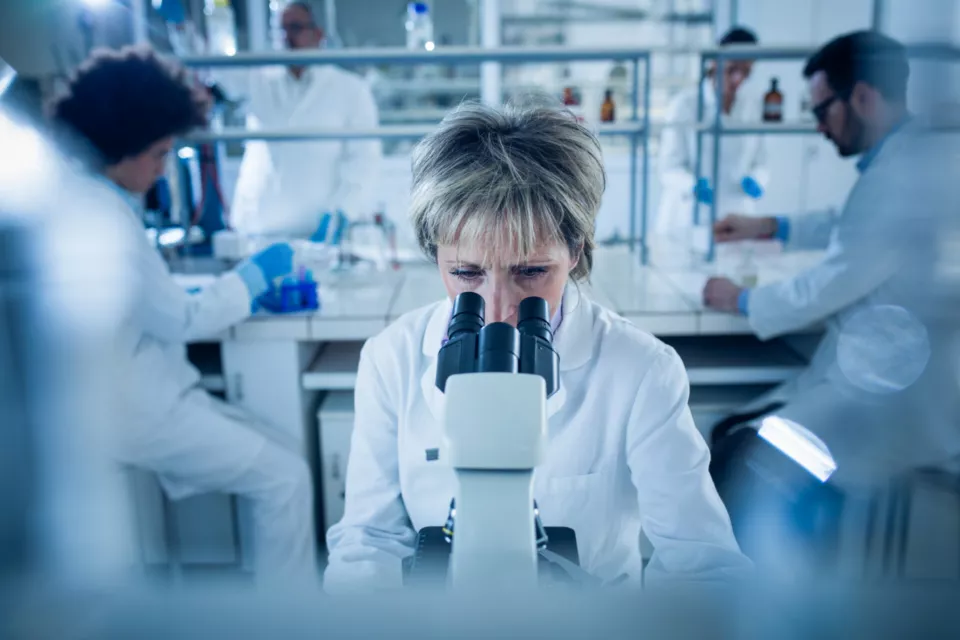 Senior forensic scientist analyzing something through a microscope in laboratory. There are people in the background.